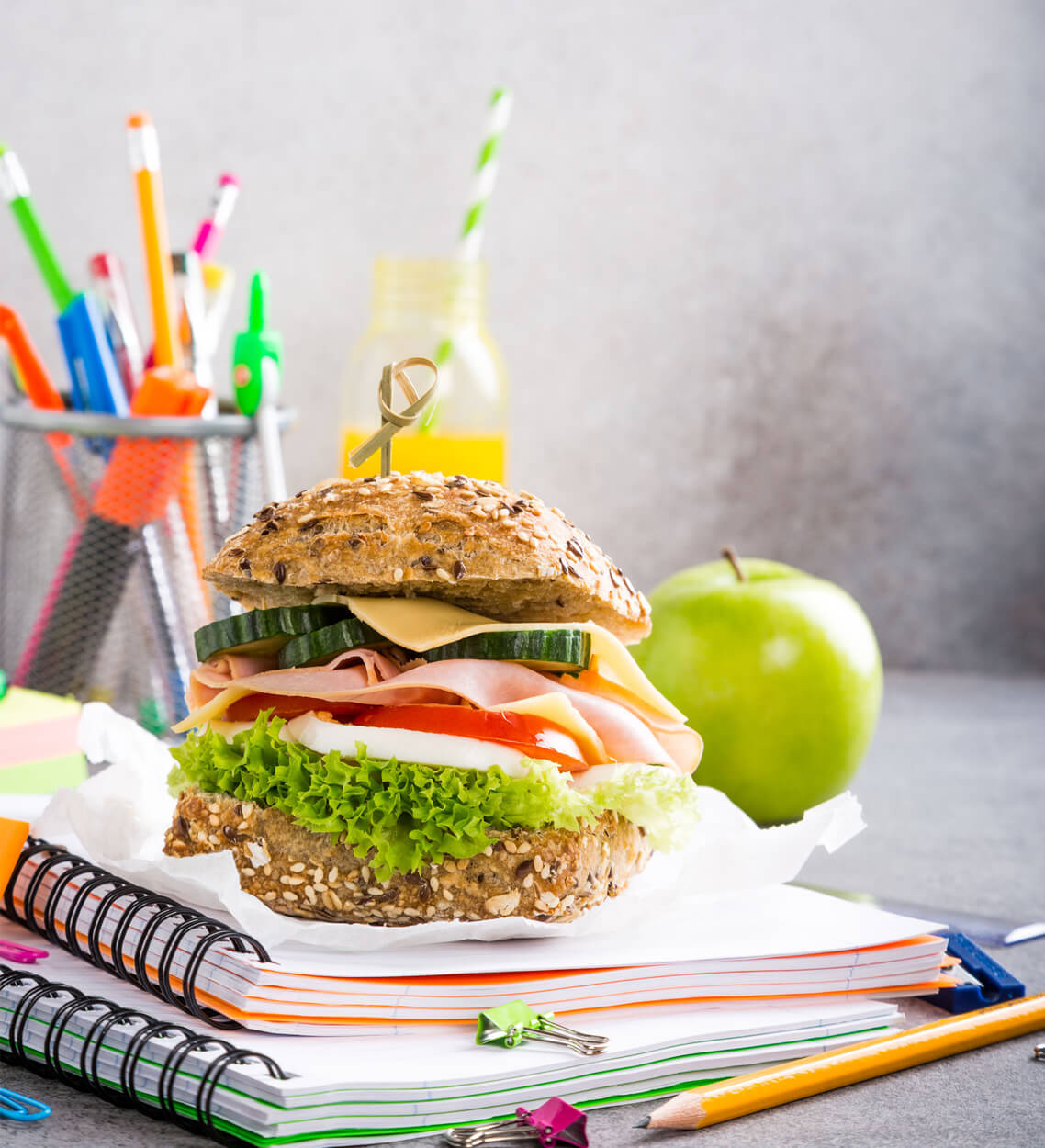 Metro Canteens Adelaide, Canteen Suppliers in Adelaide of a school lunch - salad roll sitting on school books with a green apple and pencils in the background.