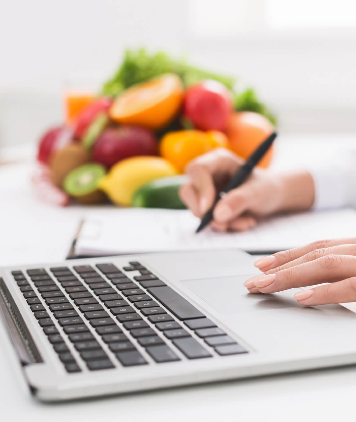 Metro Canteens Adelaide, canteen service providers image of a lady working on a laptop computer while writing a list on a paper notepad with a stack of fresh fruit blurred in the background.