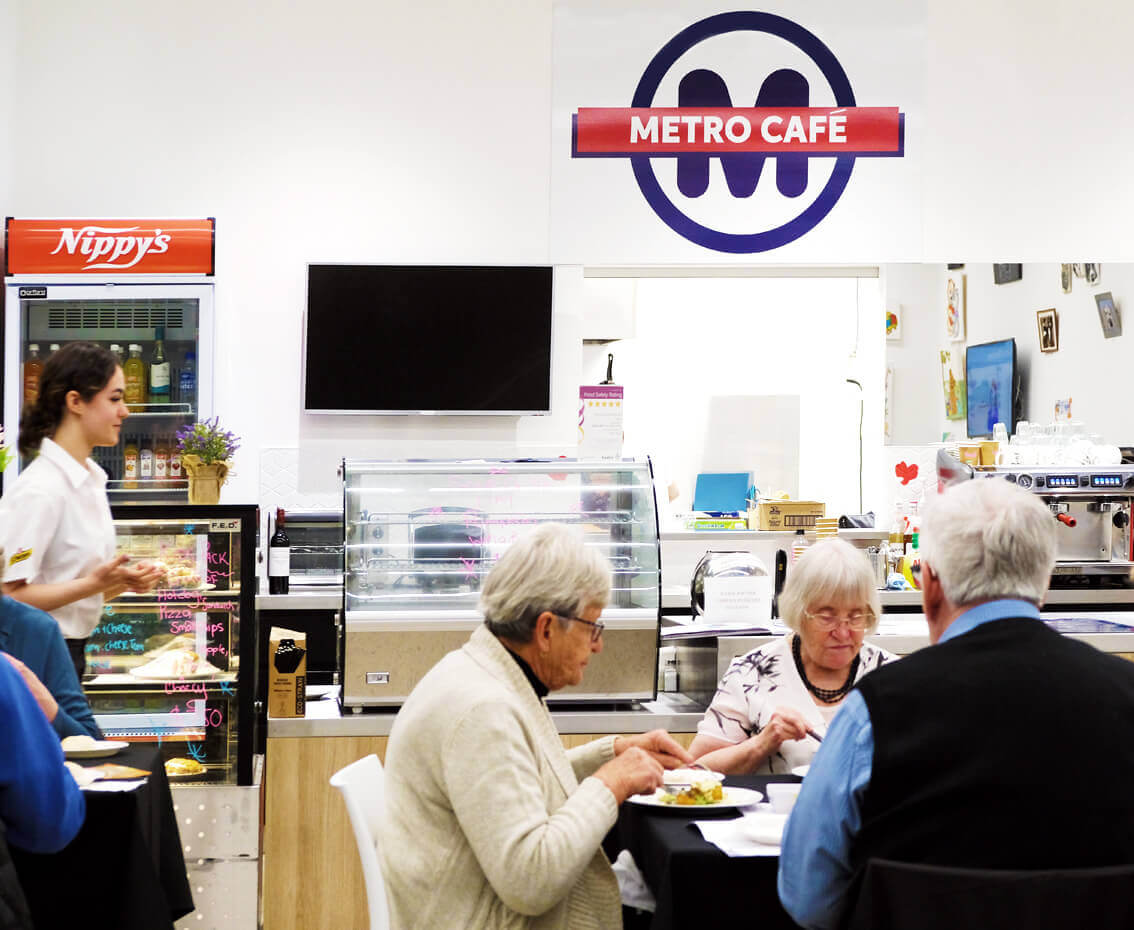 Metro Canteens - Commercial Canteen Outsourcing page photo of 3 elderly people eating at a Metro Cafe with a young waitress walking past.