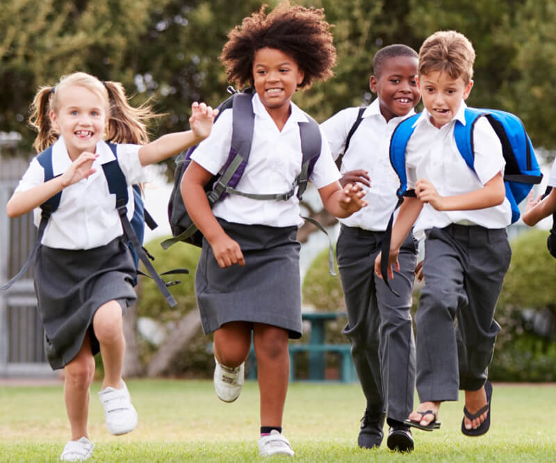 Metro Canteens Adelaide, Canteen Suppliers in Adelaide image of school four children running towards the camera, across a grass oval with school bags on their backs.